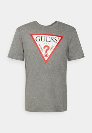 Camiseta de algodón gris con mangas cortas y cuello redondo. Presenta un logo en forma de triángulo rojo con la palabra "GUESS" y texto adicional en su interior.