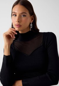 Junge Frau mit blauen Augen und langen braunen Haaren trägt ein schwarzes durchsichtiges Oberteil mit Strasssteinen und große, baumelnde Kristallohrringe, hält ihre Hand nahe am Gesicht.