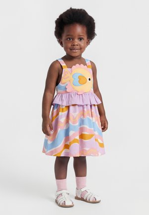 Småbarn iført en farverig kjole med bølgede mønstre og et stort fiskedesign, kombineret med lyserøde sokker og hvide sandaler.