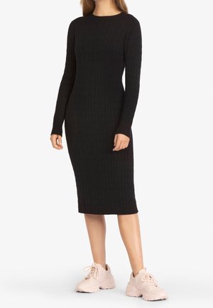 Kobieta w dopasowanej, długorękawowej, czarnej sukience midi z grubym splotem, nosząca jasnoróżowe, masywne sportowe buty, stojąca na białym tle.