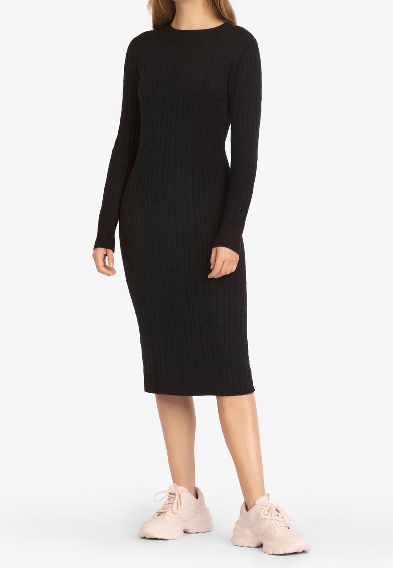 Femme portant une robe midi ajustée à manches longues en maille torsadée noire, avec des baskets roses claires épaisses, debout devant un fond blanc.