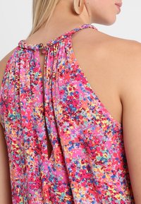 Haut sans manches à fleurs présentant des teintes roses vives, des motifs multicolores, un design à ouverture clé au dos et un col froncé avec des détails décoratifs.