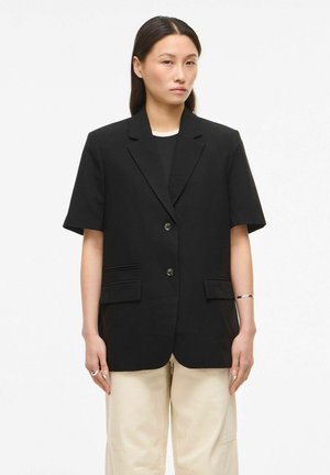 Ženska s ravnimi temnimi lasmi, ki nosi črni blazer s kratkimi rokavi, črno majico in bež hlače, stoji pred belim ozadjem.