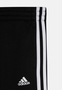 Pantalones deportivos negros con tres franjas blancas a los lados, con una textura de tela suave y un logotipo de Adidas bordado en blanco.