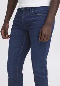 Donkerblauwe denim jeans met een slim fit, voorzien van riemlussen, voorzakken en een gladde textuur. De stof is rekbaar en heeft een lichte textuur.