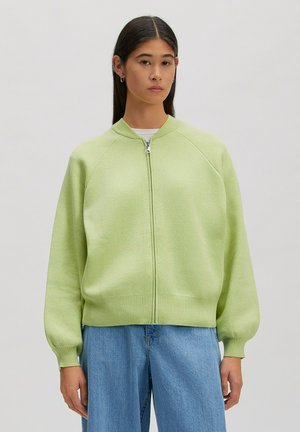 Vrouw met lang donker haar draagt een lichtgroene trui met rits en losse blauwe jeans, staande tegen een effen lichte achtergrond.