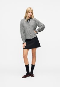 Femme blonde debout, main sur la hanche, portant un chemisier à carreaux noir et blanc, une jupe noire, des chaussettes noires et des ballerines marron, sur fond blanc.