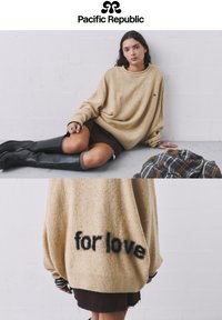 Pulover oversize bej din tricot, cu un finisaj texturat, manșete cu rib și inscriptionat pe spate cu "pentru iubire". Fustă maro și cizme negre înalte.