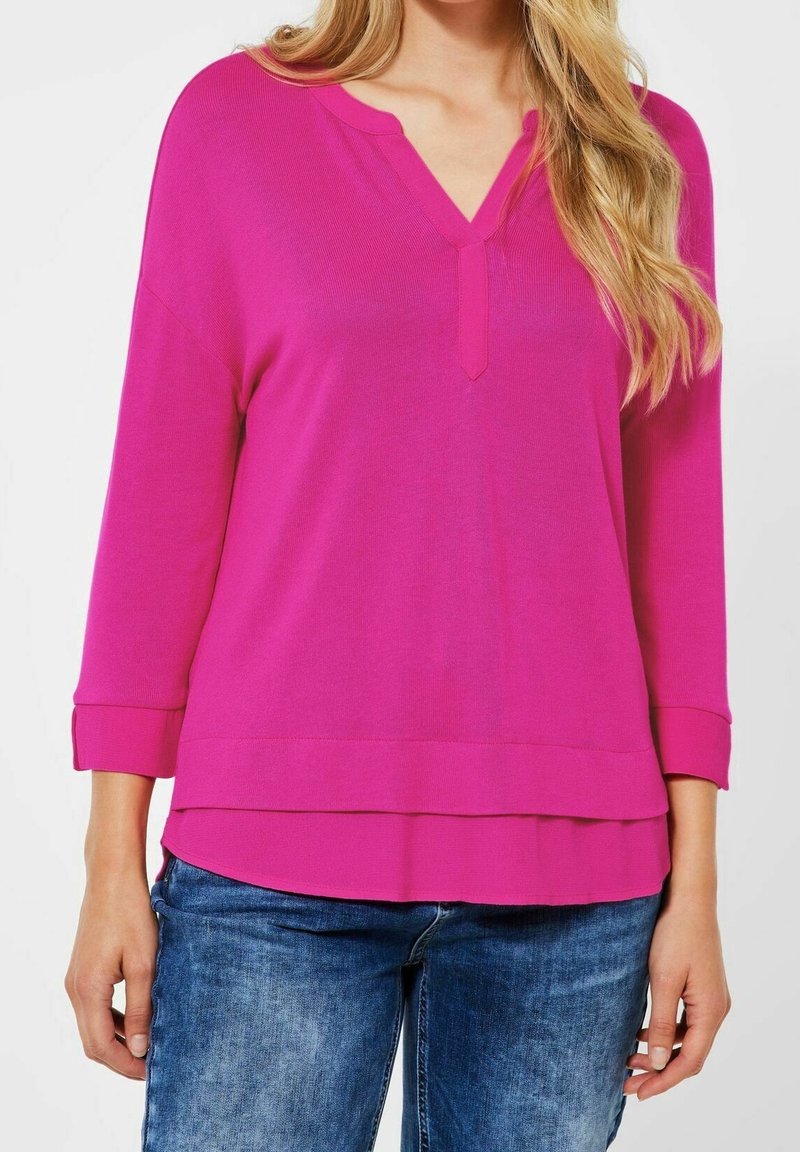 Femme portant un top à manches longues rose vif avec une encolure en V et un ourlet superposé, associé à un jean en denim bleu.