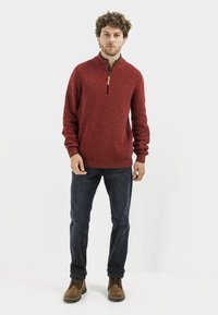 Sweater zippé rouge en tricot avec un tissu texturé, doté d'un col haut et d'un tirette en bois brun. Associé à un jean foncé et des bottes brunes.