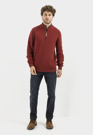 Red knit zip-up sweater with textured fabric, featuring a high collar and a brown wooden zipper pull. Paired with dark jeans and brown boots.