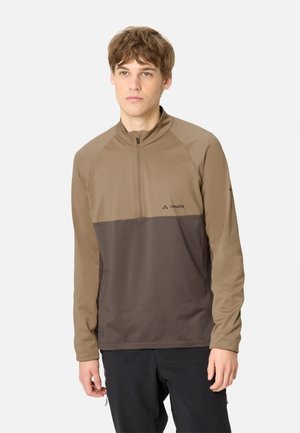 Giovane uomo che indossa un maglione marrone a due tonalità con maniche lunghe e cerniera, in piedi davanti a uno sfondo chiaro e semplice.
