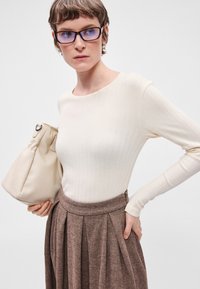 Krém színű bordás, hosszú ujjú blúz barna halszálkás redőzött szoknyával párosítva, bézs, puha bőrtáskával a kezében. Átlátszó keretes szemüveg van rajta.