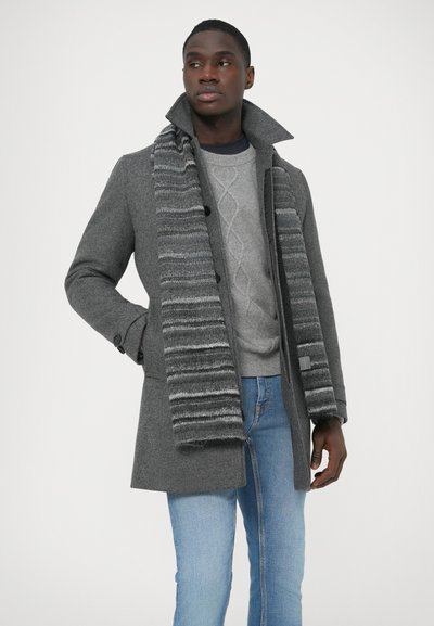 Γκρι μάλλινο παλτό με ψηλό γιακά, μαύρα κουμπιά, φορεμένο πάνω από ανοιχτό γκρι πλεκτό πουλόβερ. Τυλιγμένο με ριγέ γκρι κασκόλ, σε συνδυασμό με μπλε τζιν.