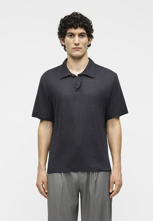 Άνδρας με σγουρά σκούρα μαλλιά στέκεται στραμμένος μπροστά φορώντας ένα χαλαρό μαύρο κοντομάνικο πόλο πουκάμισο και γκρι πλισέ παντελόνια.