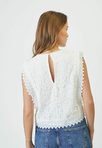 Top bianco in pizzo con bordi scallop e dettaglio apertura a chiave sul retro. Abbinato a pantaloni shorts in denim, che mostrano un motivo floreale testurizzato.