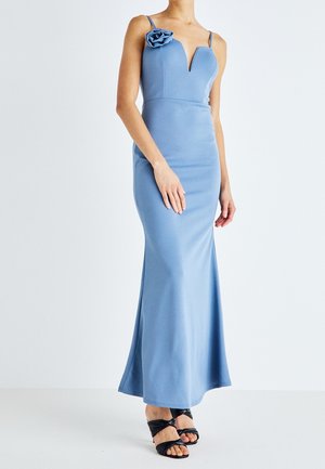 Kobieta ubrana w dopasowaną, sięgającą podłogi jasnoniebieską suknię na cienkich ramiączkach z kwiatowym zdobieniem na lewym ramieniu, w połączeniu z czarnymi sandałami na obcasie.