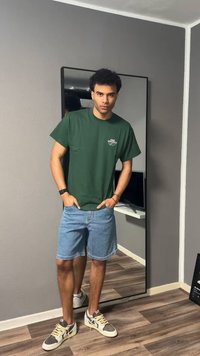 Jaunas vyras su garbanotais plaukais, dėvintis žalią Nike Athletics marškinėlį, džinsinius šortus ir sportbačius, stovintis priešais viso ūgio veidrodį kambaryje.