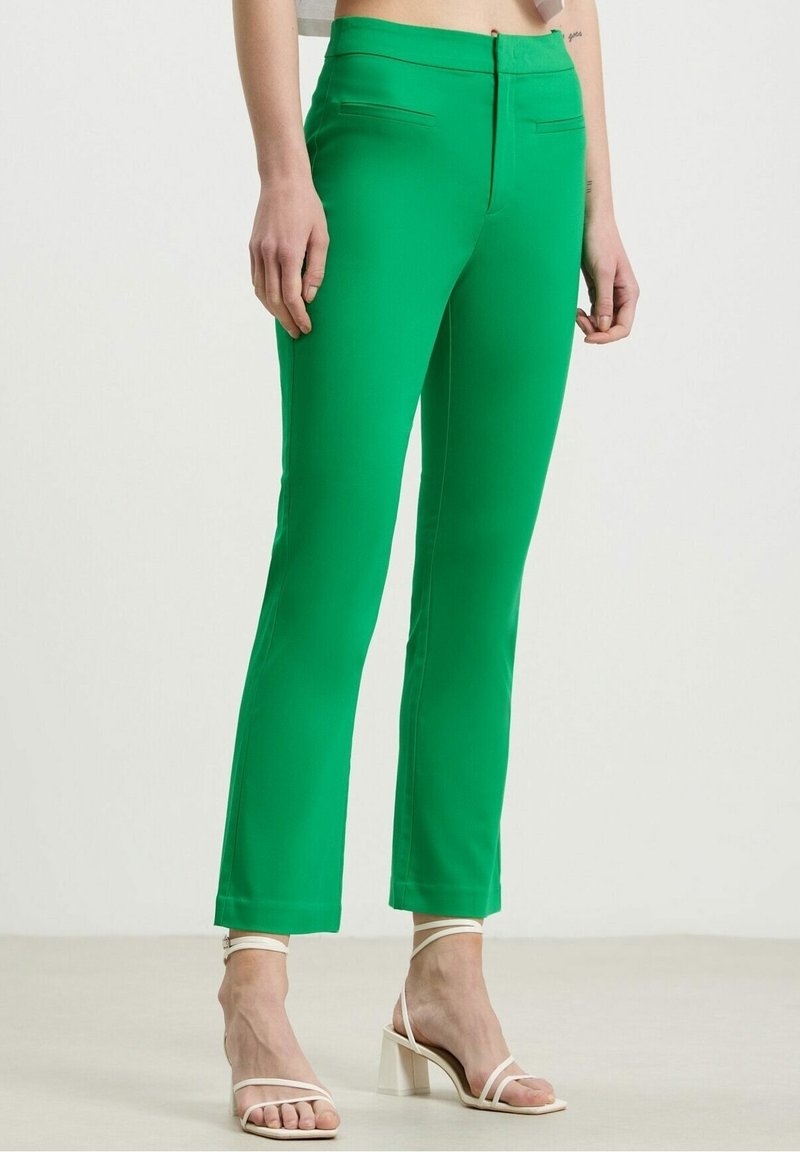 Pantalon slim vert vif longueur cheville porté avec des sandales blanches à talons blocs et à brides sur une personne debout.