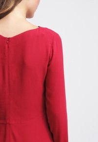 Punainen mekko, jossa on pitkät hihat, sileä pinta ja avainreikäkietaisusuljin selässä. Yksinkertainen muotoilu ilman koristeluja tai kuvioita.