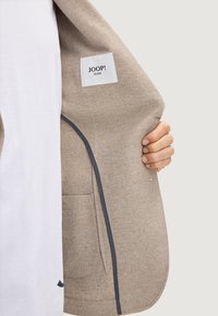 Blazer beige con motivo a spina di pesce. L'interno presenta un'etichetta contrassegnata "JOOP! JEANS" e un bordo scuro a contrasto lungo il margine.