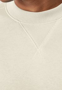 Sudadera de color beige claro con textura, cuello redondo acanalado y detalle de costura en forma triangular en el pecho. Acabado de tela suave.