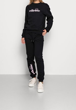 Person iført en sort ellesse sweatshirt og matchende joggingbukser med pink ellesse-logo, kombineret med hvide sneakers, stående mod en ensfarvet baggrund.