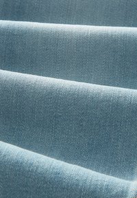 Šviesiai mėlyna džinsinė medžiaga su tekstūruota paviršiumi, turinti matomas įstrižas audimo raštus ir sulankstytas dalis, išryškinančias sklandžias linijas ir spalvą.