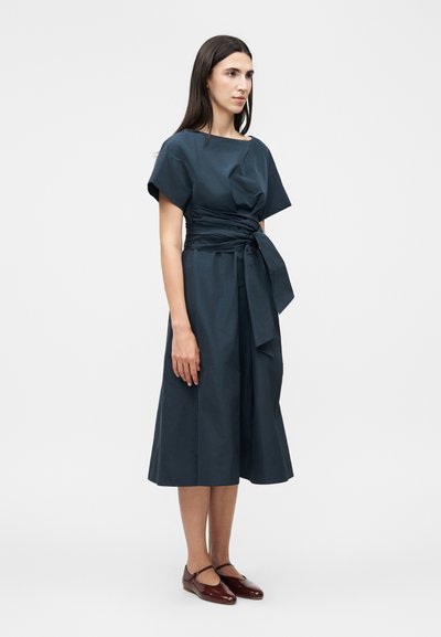 Γυναίκα με μακριά σκουρόχρωμα μαλλιά που φοράει ένα σκούρο μπλε midi φόρεμα με φαρδιά ζώνη δεμένη στη μέση και σκούρα καφέ παπούτσια Mary Jane, στέκεται πλαγίως.