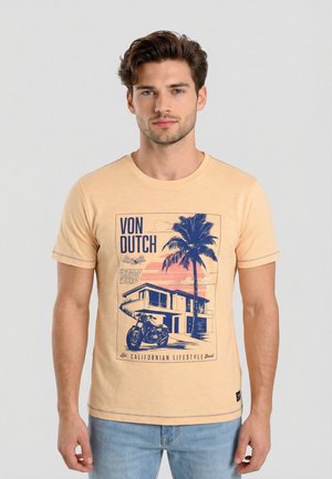 Joven con camiseta beige con un gráfico azul de una casa moderna, palmera, motocicleta y texto "VON DUTCH" y "Estilo de vida californiano."