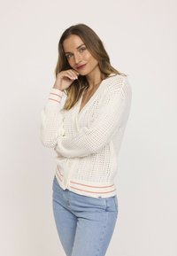 Cardigan blanc tricoté avec une trame ouverte, décolleté en V et boutons. Les poignets et l'ourlet côtelés présentent des rayures orange. Assorti à un jean bleu clair.