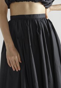 Falda negra plisada hecha de tela texturizada; presenta una cinturilla ajustada y un suave drapeado. Mano descansando sobre la tela de la falda.