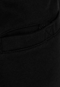 Schwarzer Baumwollstoff mit strukturierter Oberfläche, der ein kleines Taschendetail und Nähakzente aufweist und Teil eines Kleidungsstücks ist.