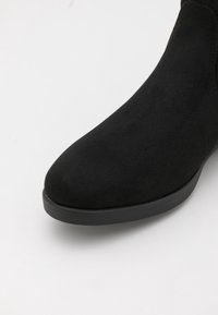 Black suede ankle boot with a rounded toe, flat black sole, and minimal stitching detail along the upper edge. Smooth texture.