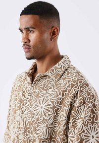 Camisa marrón claro con bordado floral y abstracto en blanco, con cuello y cierre de botones en la parte delantera. Detalle de tejido texturizado visible.