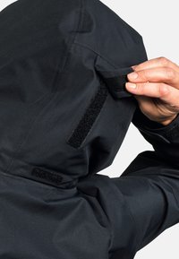 Händisches Anpassen der Kapuze einer schwarzen Jacke mit Klettverschlussriemen auf einem glatt strukturierten Stoff.