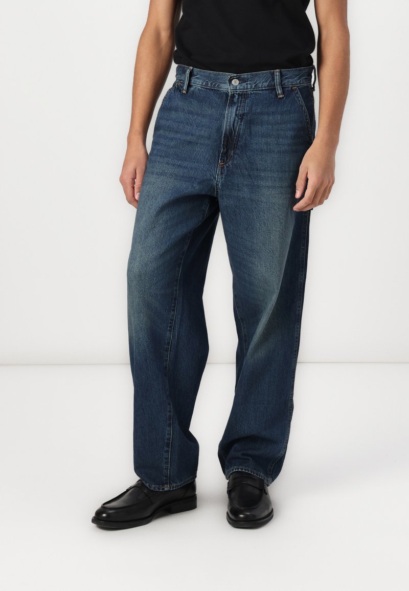 Mørkeblå denimjeans med løs passform, rett ben og synlig søm. Kombinert med sorte sko og en svart skjorte.
