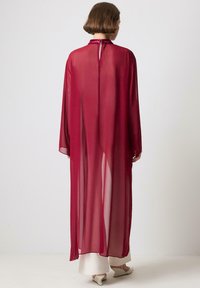 Długi, przejrzysty bordowy kimono z szerokimi rękawami, wysokim kołnierzem i detalem w formie kluczyka z tyłu, noszony na białym stroju.