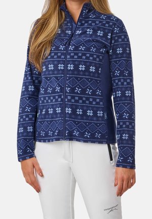 Kobieta ubrana w granatową polarową kurtkę na zamek błyskawiczny z jasnoniebieskimi wzorami płatków śniegu oraz białe spodnie z czarną kieszenią na zamek.