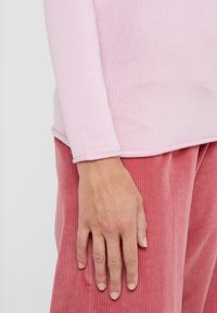 Langärmliges, hellrosa Strickoberteil mit abgerundetem Saum, kombiniert mit weichen rosa Cordhosen mit vertikalen Streifen und sichtbaren Nähten.