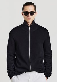 Schwarzer Reißverschluss-Pullover mit hohem Kragen, strukturiertem Strickmaterial und gerippten Bündchen. Modell trägt schwarze Sonnenbrille vor neutralem Hintergrund.