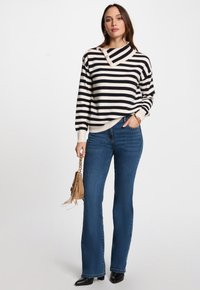 Maglione a righe con un collo alto e dettagli in bottone, abbinato a jeans blu a campana e stivali neri alla caviglia. La modella tiene una borsa con frange.