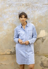 Jeune femme aux cheveux bouclés portant une robe en jean bleu clair boutonnée, debout contre un mur texturé et usé.