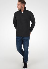 Dunkelgrauer, strukturiertes Zopfstrickpullover mit einem Reißverschlusskragen, kombiniert mit blauen Jeans und schwarzen Sneakers. Der Stoff hat ein texturiertes, geripptes Design.