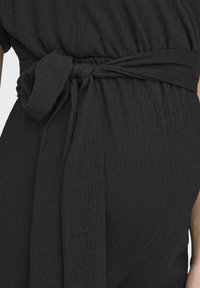 Vestido negro texturizado con tela tejida, cintura ajustada con un cinturón atado y patrones sutiles de pliegues.