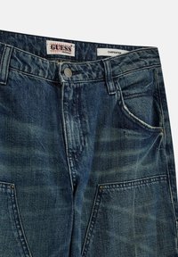 Dunkelblaue Zimmermannsjeans mit verblassten Bereichen, Knopfverschluss, mehreren Taschen und sichtbarem Markenlabel "Guess Originals" im Bund.