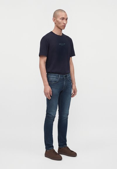 Άνδρας που στέκεται φορώντας μαύρο T-shirt Dondup, εφαρμοστά μπλε τζιν και καφέ αθλητικά παπούτσια σε απλό ανοιχτό φόντο.