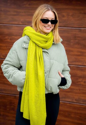 Groene corduroy pufferjack, zwart langemouwtop en een felgele sjaal die tegen een bruine houten achtergrond zijn weergegeven.