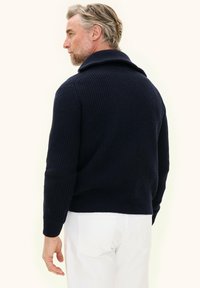 Uomo con capelli e barba grigi, vestito con un maglione a coste blu scuro e pantaloni bianchi, voltato leggermente di lato davanti a uno sfondo neutro.