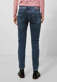 Jeans de corte slim en azul oscuro, con una textura desgastada, dos bolsillos traseros y una etiqueta de marca en la cintura. Combinados con botas de tobillo negras.